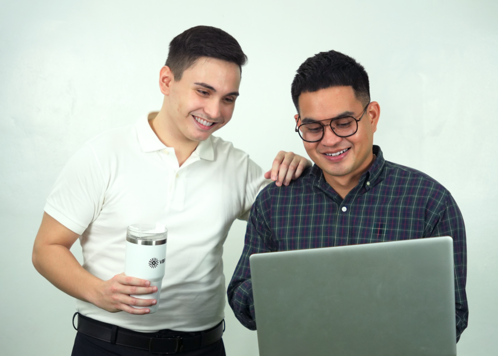 Two male team members looking at the laptop, male at the right is holding the laptop while man at the left is holding a VBP thermal cup 